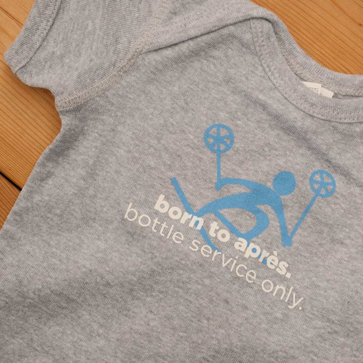 Grey baby onesie with a blue skier graphic and ‘born to après. bottle service only’ lettering.
