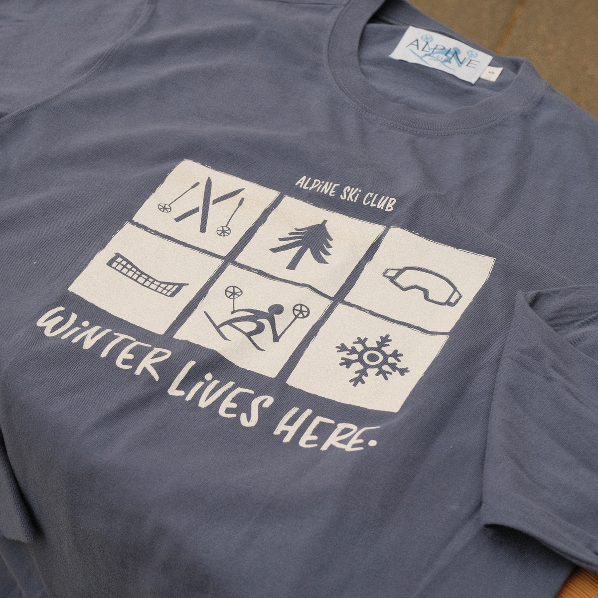 Washed royal blue Alpine Ski Club adult long-sleeve tee with six winter icons and ‘Winter Lives Here’ lettering.