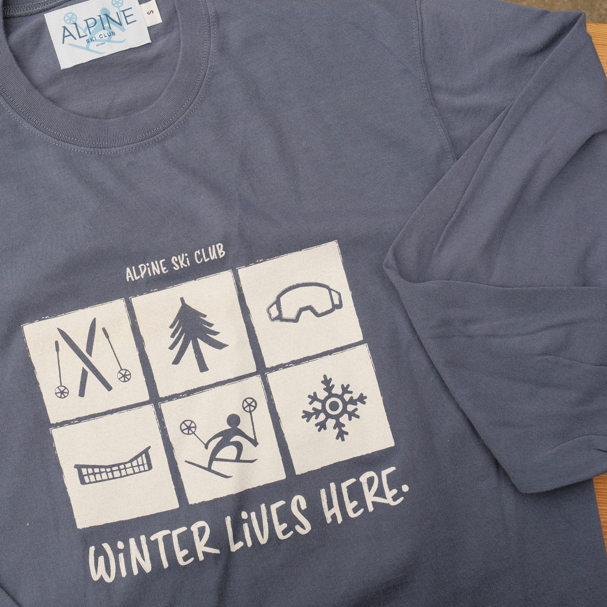 Close-up of the ‘Winter Lives Here’ graphic on a washed royal blue Alpine Ski Club tee, with long sleeve folded across.