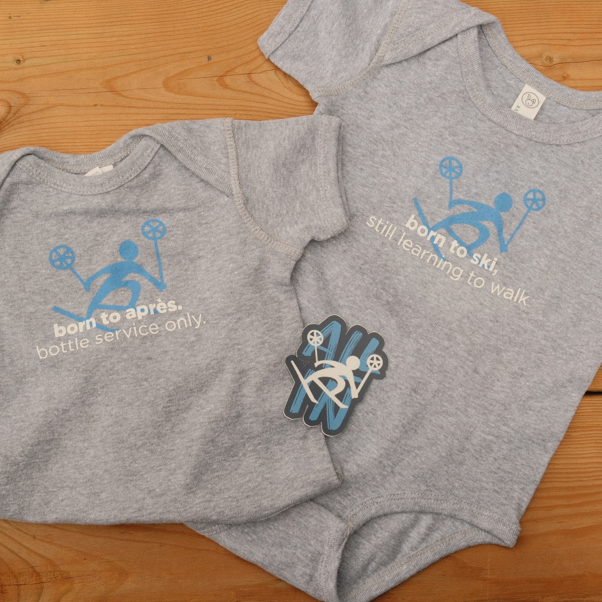 Two grey Alpine Ski Club baby onesies with blue skier graphics and playful ski and après slogans.