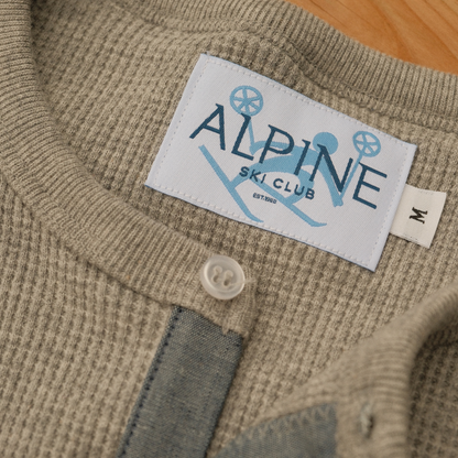 Close-up of the Alpine Ski Club neck label, size M tag and button neckline on a light grey waffle-knit Henley.