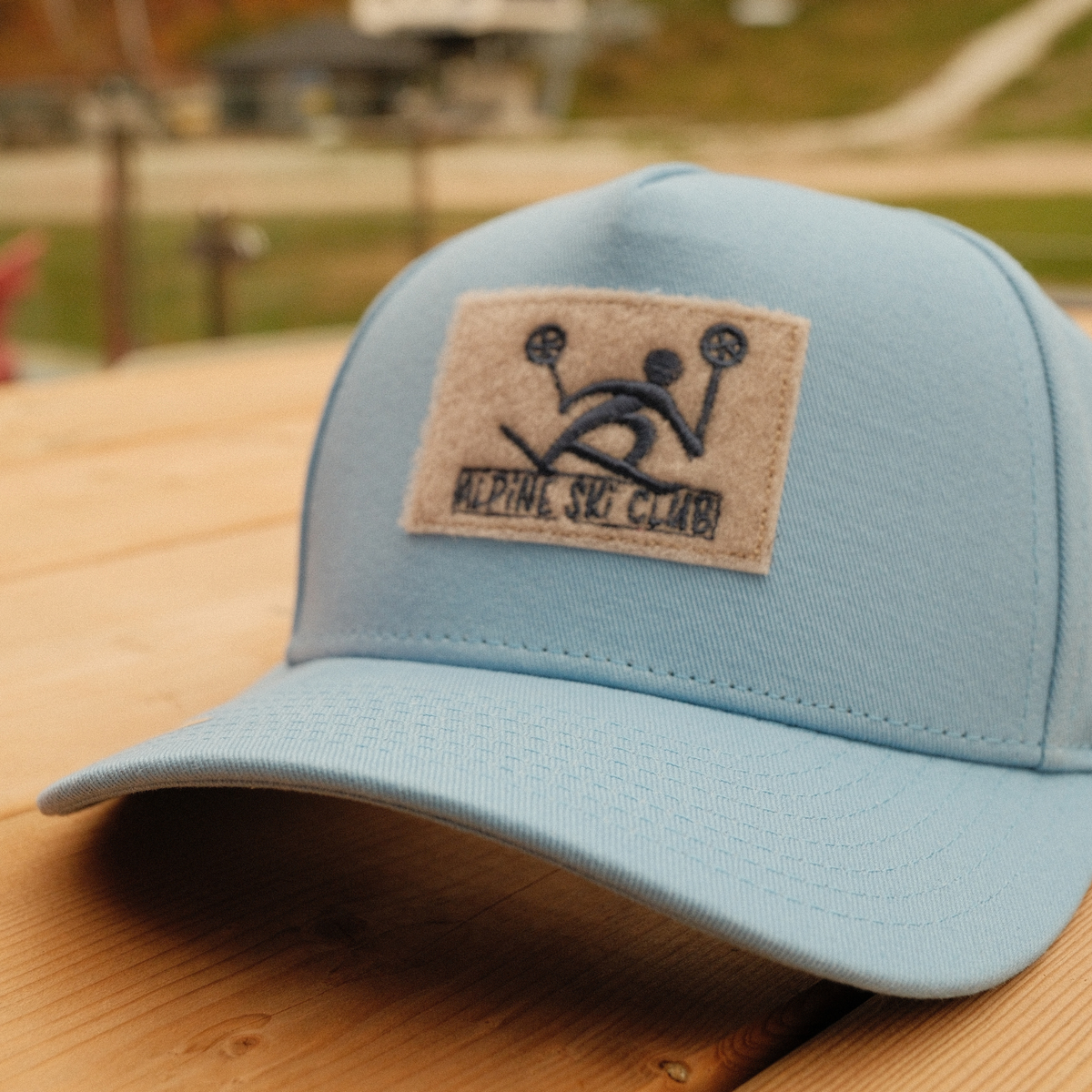Carolina blue kids’ baseball cap with a tan Alpine Ski Club skier patch, resting on a wooden table.
