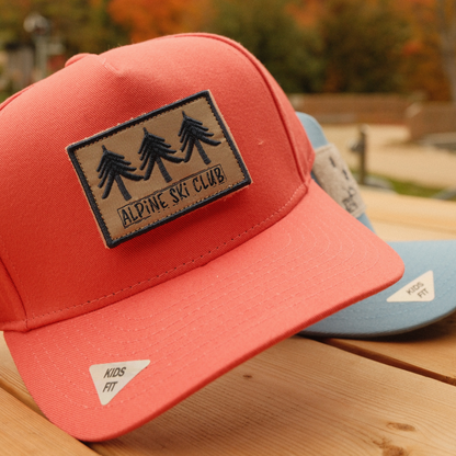 Coral kids’ baseball cap with a tan Alpine Ski Club patch featuring three trees, with a blue cap behind.