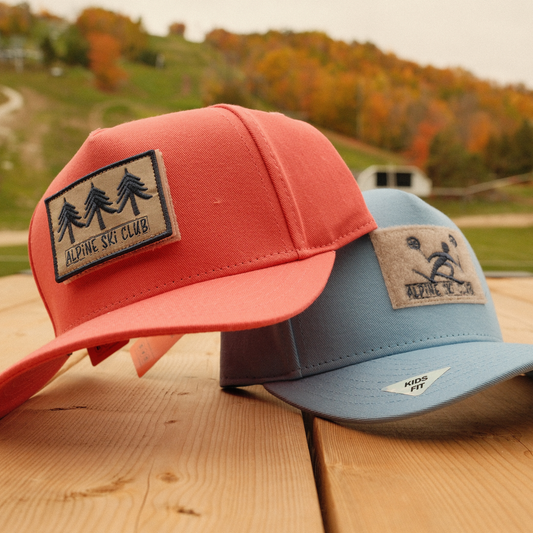 Coral and Carolina blue Alpine Ski Club kids’ caps with tree and skier patches, displayed against an autumn ski hill.