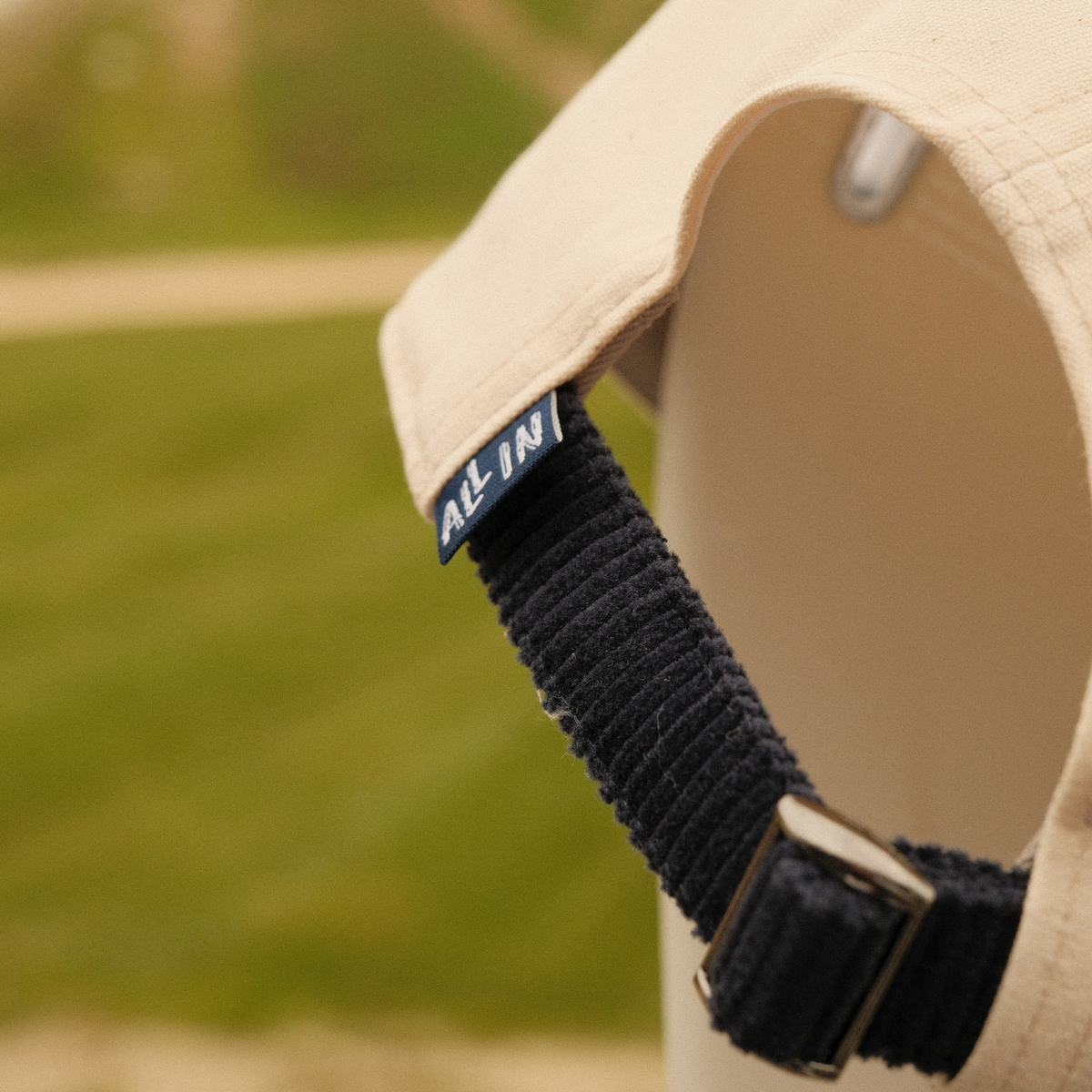 Back of cream Alpine Ski Club dad hat showing a navy adjustable strap, metal buckle and ‘ALL IN’ tag.