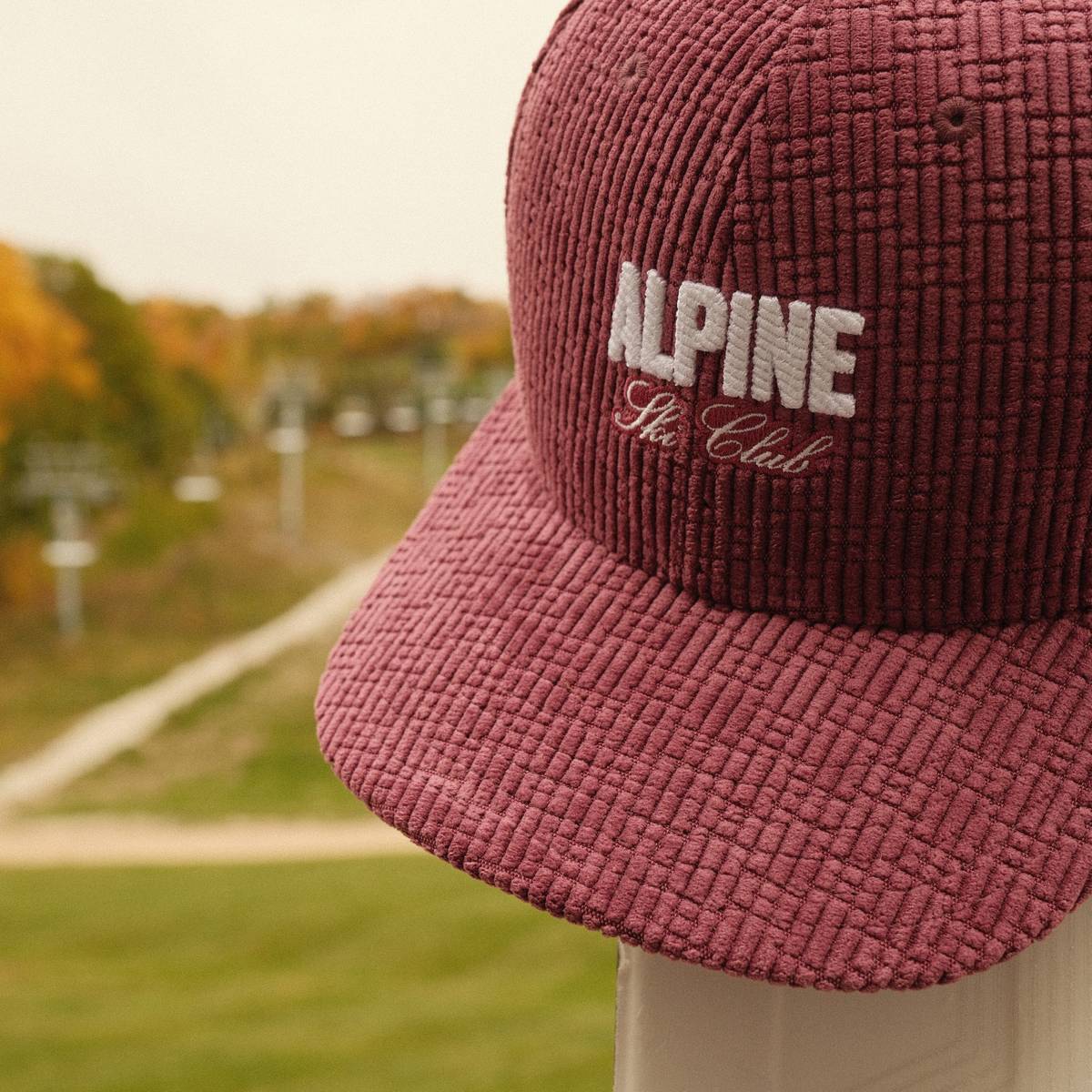 Plum corduroy Alpine Ski Club dad hat with white embroidered lettering against an autumn ski hill.