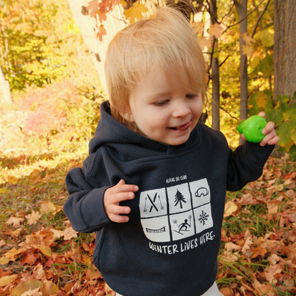 Toddler wearing a navy Alpine Ski Club ‘Winter Lives Here’ hoodie outdoors among autumn leaves.